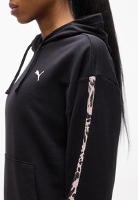 Γυναίκα που φοράει μαύρο φούτερ με κουκούλα Puma με κορδόνια, τσέπη καγκουρό και λωρίδα με λεοπάρ σχέδιο στο μανίκι, εμφανιζόμενη από το πλάι με φόντο το λευκό.