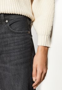 Jean en denim noir à coupe ajustée, présentant une texture subtile, un devant avec des boutons et une poche latérale, assorti à un pull crème côtelé.