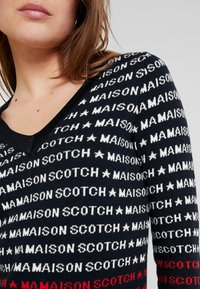Sweter wykonany na drutach z dekoltem w serek, czarną bazą i powtarzającym się tekstem w białym i czerwonym kolorze, z akcentami w postaci gwiazd. Miękka tekstura i dopasowany krój.