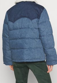 Personne portant une veste en jean rembourrée bleue avec un empiècement bleu marine et un col en Sherpa blanc, accompagnée de pantalons vert foncé.