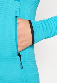 Κοντινό πλάνο ενός μακρυμάνικου τοπ σε τιρκουάζ χρώμα με υφή, που διαθέτει τσέπη με φερμουάρ και μαύρες λεπτομέρειες στο μανίκι.