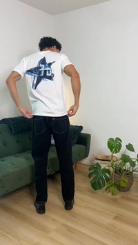 Άνδρας που προσαρμόζει λευκό T-shirt με μπλε αστέρι στο πίσω μέρος, όρθιος κοντά σε πράσινο καναπέ και μεγάλο φυτεμένο φυτό σε φωτεινό δωμάτιο.