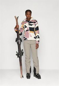 Donna con scarponi da sci che tiene gli sci, indossa pantaloni beige e una giacca in pile con motivo multicolore, in piedi su uno sfondo neutro.