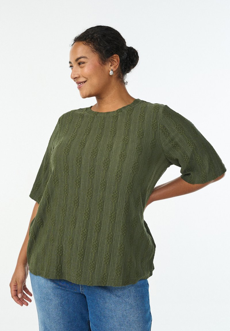 Zelený top s texturou a vlnitým vzorem, krátkými rukávy a zaobleným výstřihem, ve spojení s riflemi s vysokým pasem.