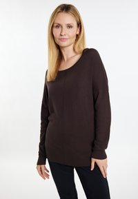 Braun gestrickter Pullover mit weitem Rundhalsausschnitt, langen Ärmeln, gerippten Bündchen und einem etwas längeren Rückensaum, kombiniert mit dunklen Hosen.