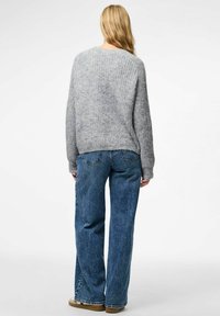 Sieviete ar gariem blondiem matiem, tērpusies pelēkā teksturētā džemperī, zils platās kājas biksēs un brūnās kurpēs, stāv ar muguru pret baltu fonu.