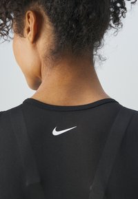 Črna športna majica z okroglim izrezom, na hrbtni strani pa ima belo Nike logotip. Tkanina se zdi gladka s subtilno teksturo.