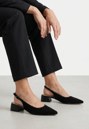 Person trägt schwarze Slingback-Schuhe mit spitzer Kappe und niedrigem Blockabsatz sowie kurze schwarze Hose, sitzt auf hellem Boden.
