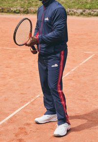 Marinträningsdräkt med röda sidostreck, jacka med dragkedja och matchande byxor. Vita tennis skor och ett tennisrack hålls i händerna. Bakgrund av lera.