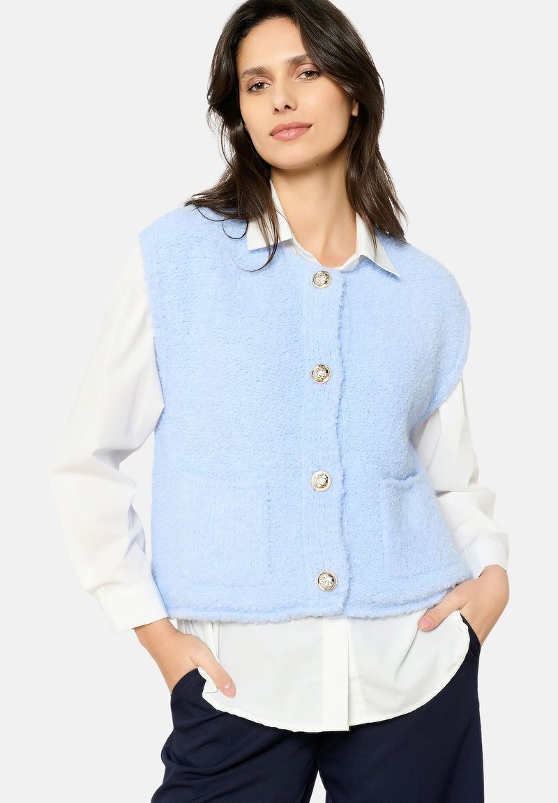 Gaiši zils bezpiedurkņu veste, izgatavota no teksturēta auduma, ar lielām sudraba pogām un divām priekšējām kabatām, uzvilkta virs balta krekla.