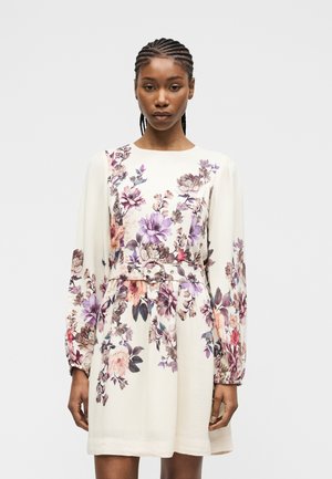 Femme portant une robe blanche à manches longues avec un imprimé floral en violet, rose et marron, avec une ceinture en tissu assortie à la taille.