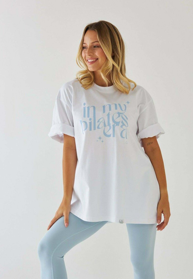 Biała, oversize'owa koszulka z niebieskim napisem "w mojej erze pilatesu", podwinięte rękawy, w połączeniu z jasnoniebieskimi legginsami, na neutralnym tle.