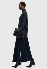 Navy turtlenecksweater, vide benbukser i navy, sorte ankelstøvler med guldaccenter, holder en sort clutcheskabel. Ensfarvet baggrund.