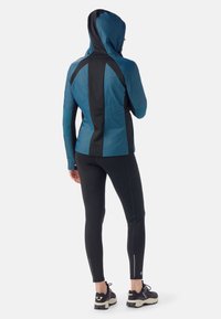 Gekapuzeerde jas in teal en zwart, met een strakke uitstraling, rekbaar materiaal en een getailleerde pasvorm. Gecombineerd met zwarte leggings en sneakers.