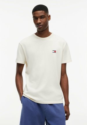 Jeune homme portant un t-shirt uni blanc cassé avec un petit logo et un short bleu texturé, debout devant un fond blanc.