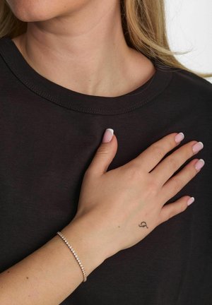 Ezüst karkötő átlátszó kövekkel, világos bőrű kézen rózsaszín körmökkel, sötét pólón nyugszik.