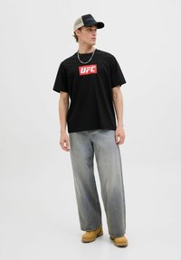 Sort kortærmet t-shirt med hvid UFC-logo, parret med løstsiddende lysegrå jeans og brune arbejdssko, under en sort og grå kasket.