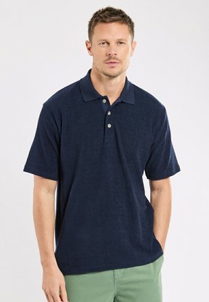 Homme portant un polo bleu marine à manches courtes et un pantalon vert clair, debout devant un fond blanc uni.