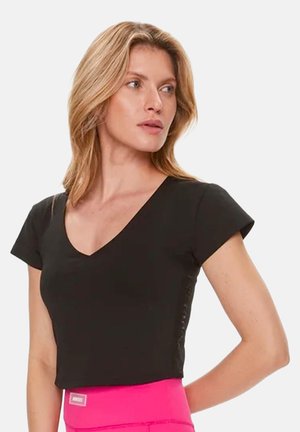 Vaaleahiuksinen nainen, jolla on musta V-kaula-aukollinen crop top ja kirkkaanpinkit korkeavyötäröiset housut, katsoo vasemmalle käsi selän takana.