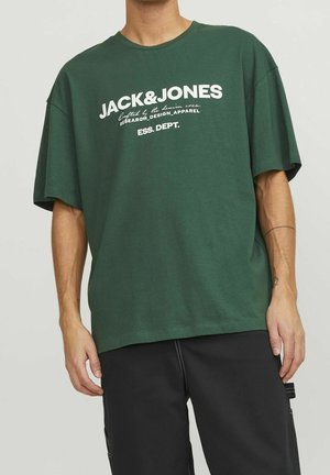 Homme portant un t-shirt vert Jack & Jones avec un texte blanc et un pantalon noir, debout bras détendus contre un fond clair uni.
