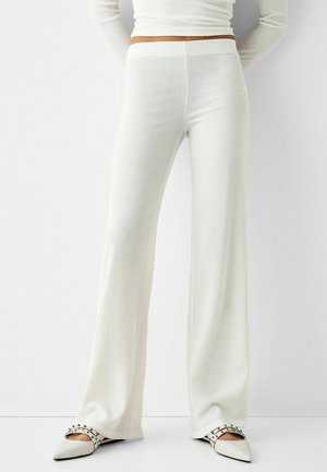 Person iført hvide, højtaljede bukser med vidt ben og tekstur samt hvide spidse flade sko med sorte nitter, stående foran en ensfarvet hvid baggrund.