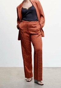 Un ensemble deux pièces en tissu de couleur rouille et à motif. Pantalons longs à jambes droites et blazer ajusté avec un motif de petits points subtils. Camisole noire en dessous.