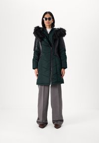 Puffy green down coat with a detachable black faux fur hood, quilted body, and black ribbed sleeves, paired with gray wide-leg trousers.