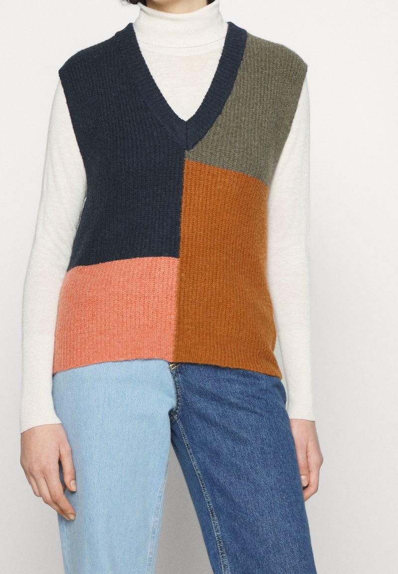 Veste tricotée sans manches à col en V avec sections bleu marine, olive, rouille et corail, portée sur un col roulé blanc et un jean bicolore bleu.