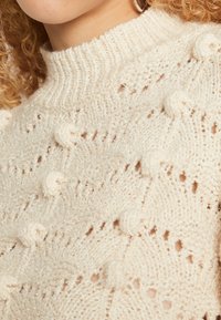 Cremefarbener Strickpullover mit hohem geripptem Kragen, der durchgängige strukturierte Muster und Bobbelakzente aufweist. Leicht und weiche Textur.