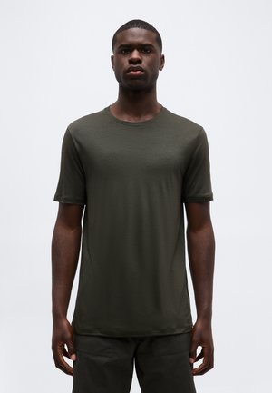Homme adulte debout, portant un t-shirt à manches courtes olive à col rond et un pantalon foncé, devant un fond clair uni.