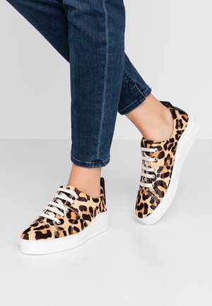 Ben iført mørkeblå jeans og leopardprint sneakers med hvide såler og snørebånd, stående på en lys grå gulv.