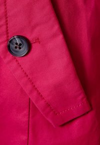 Ljust rosa tyg med en slät textur, prytt med en svart knapp med fyra hål och kontrasterande röd sömnad nära kanten.