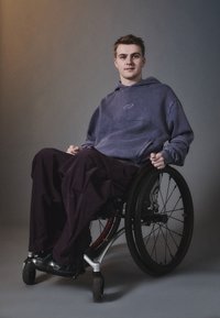 Lilla overdimensjonert hettegenser med spiraldesign, kombinert med løstsittende mørke bukser. Person som sitter i en svart rullestol med sølvfarger.
