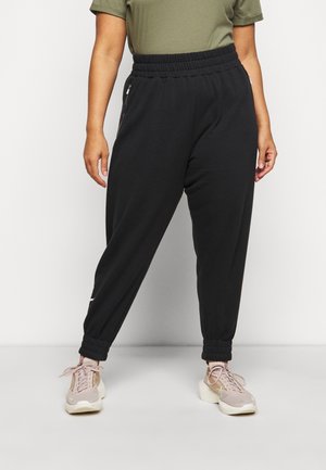 Personne portant un pantalon de jogging noir à taille élastique avec poches latérales zippées et baskets beige, debout devant un fond uni.
