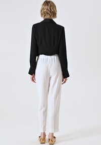 Schwarze Bluse mit langen Ärmeln, weiße maßgeschneiderte Hose mit elastischem Bund, von hinten präsentiert. Goldene Slipper mit spitzem Zeh.