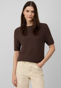 Pull marron à manches courtes, tricot texturé, col rond, forme ajustée. Associé à un pantalon beige clair. Design simple, sans motifs ni accents visibles.