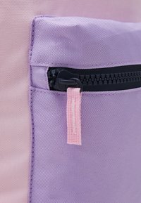 Detail batohu z fialové a růžové látky s kapsou na zip a černým zipem, na kterém je růžová látková poutko. Texturovaný, odolný materiál.