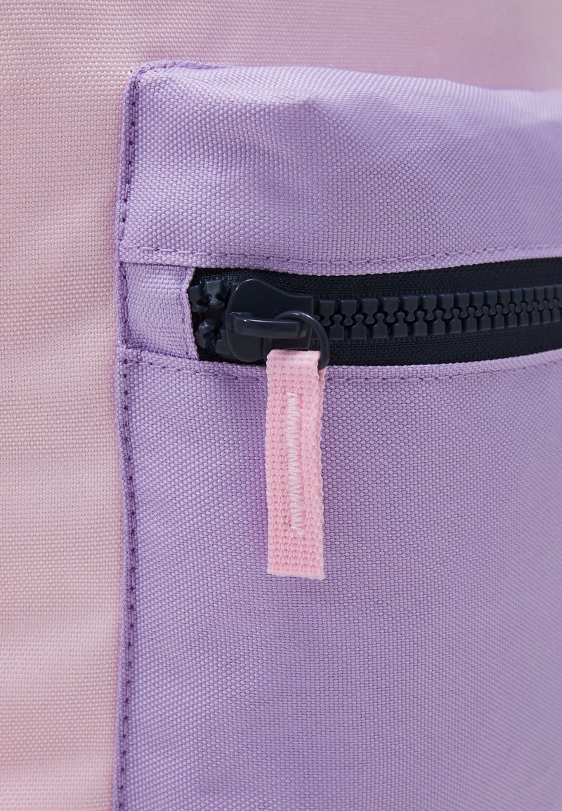 Détail d'un sac à dos en tissu violet et rose avec une poche zippée dotée d'une fermeture éclair noire et d'une tirette en tissu rose. Matériau texturé et durable.