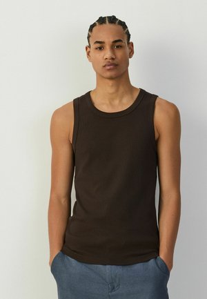 Młody mężczyzna z warkoczami, ubrany w ciemną koszulkę bez rękawów i niebieskie spodnie, stoi z rękami w kieszeniach na jednolitym tle.