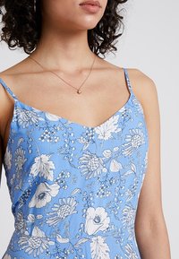 Kvinna som bär en blå blommig klänning med spagettistrol och ett fint guldhalsband med en liten berlock.