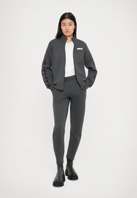 Giacca zip nera con colletto alto, abbinata a pantaloni tapered coordinati. Dettagli testurizzati in viola sulle maniche, indossata con stivali neri.