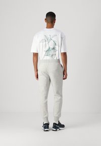 Weißes Grafik-T-Shirt mit grüner Birnenillustration auf der Rückseite, kombiniert mit hellgrauen Jogginghosen und schwarzen Sportschuhen.