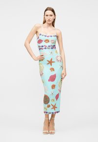 Kvinde står med hånden på hoften iført en lyseblå tætsiddende kjole med farverige mønstre af muslinger og søstjerner samt hvide højhælede sandaler.