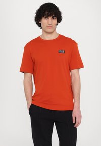 Red cotton t-shirt with a crew neckline, short sleeves, and a black logo patch on the left chest. Clean design, smooth texture.