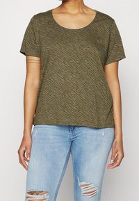 Vihreä oliivinvärinen lyhythihainen T-paita, jossa on ribbireunus ja aaltoileva rakenne sekä hienovarainen kimallus, yhdistettynä kuluneisiin vaaleansinisiin farkkuihin.