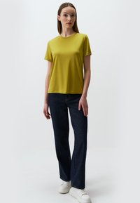 Żółta koszulka z krótkim rękawem w zestawieniu z ciemnoniebieskimi dżinsami o prostym kroju. Model nosi białe sneakersy, co podkreśla swobodny, luźny styl.