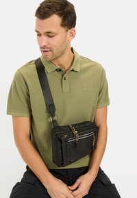 Sort nylon crossbody taske med to lynlåse, sølv hardware og en kædeaccent. Bæres over en grøn polo-shirt.