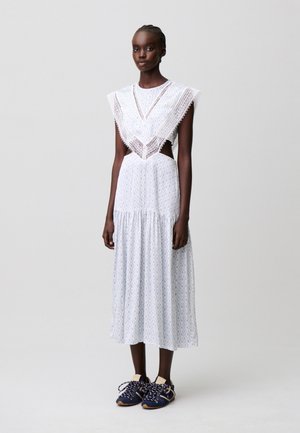 Donna snella che indossa un vestito midi bianco con rifiniture in pizzo, tagli laterali e sneakers blu navy, in piedi davanti a uno sfondo grigio uniforme.