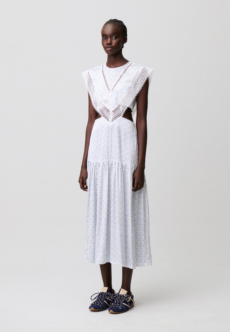 Donna snella che indossa un vestito midi bianco con rifiniture in pizzo, tagli laterali e sneakers blu navy, in piedi davanti a uno sfondo grigio uniforme.