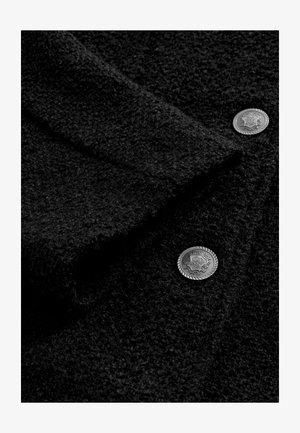 Mustaa teksturoitua kangasta, jossa on pörröinen pinta, sisältyen koristeellisia hopeanvärisiä nappeja, joissa on monimutkaisia kuvioita. Korkea kontrasti värissä ja yksityiskohdissa.